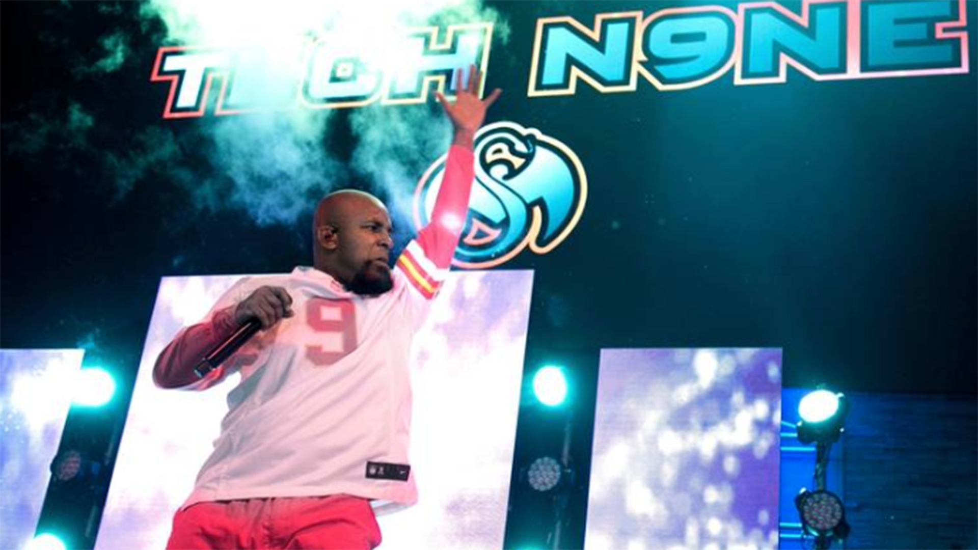 A fan raises his arms in front of a blue sign that reads "Tech N9NE" overhead.