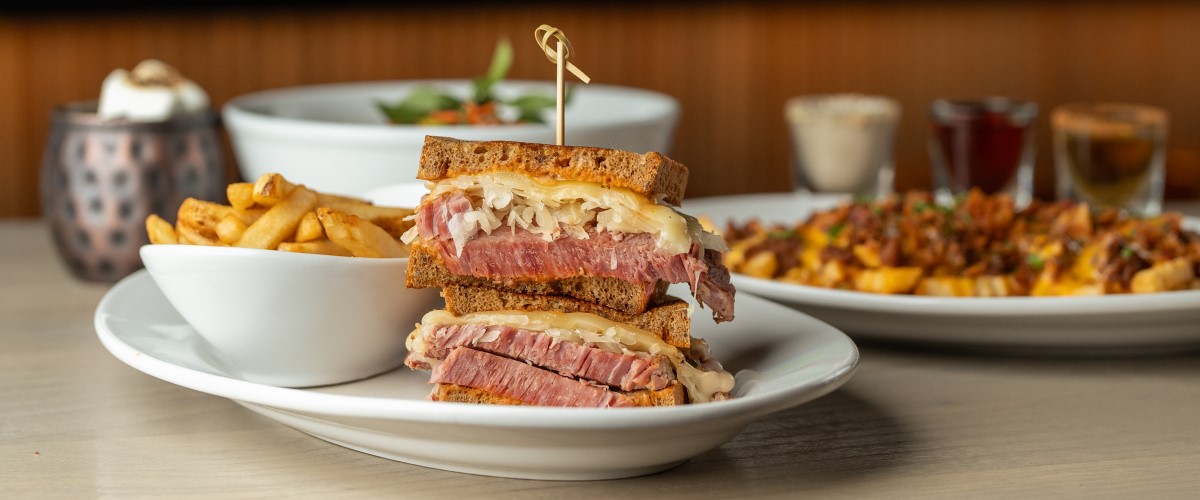 Thick-sliced corned beef Reuben with fries, pepper beef basil bowl, and loaded crinkle fries on white plates. Holiday drinks in the background - three shooters and S'mores Moscow mule.